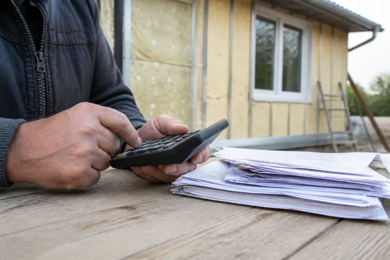 Main d'un entrepreneur tenant une calculatrice et des factures avec une maison isolée en arrière-plan.
