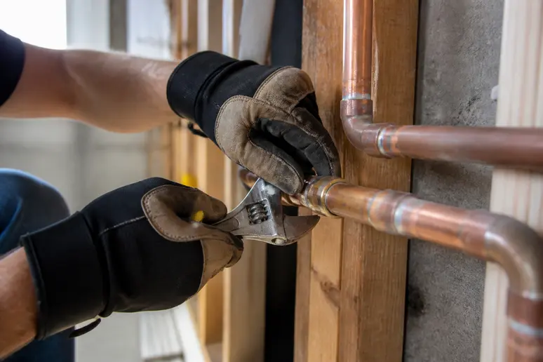 Les mains gantées d'un plombier utilisent une clé pour inspecter une conduite de cuivre dans une cave résidentielle.