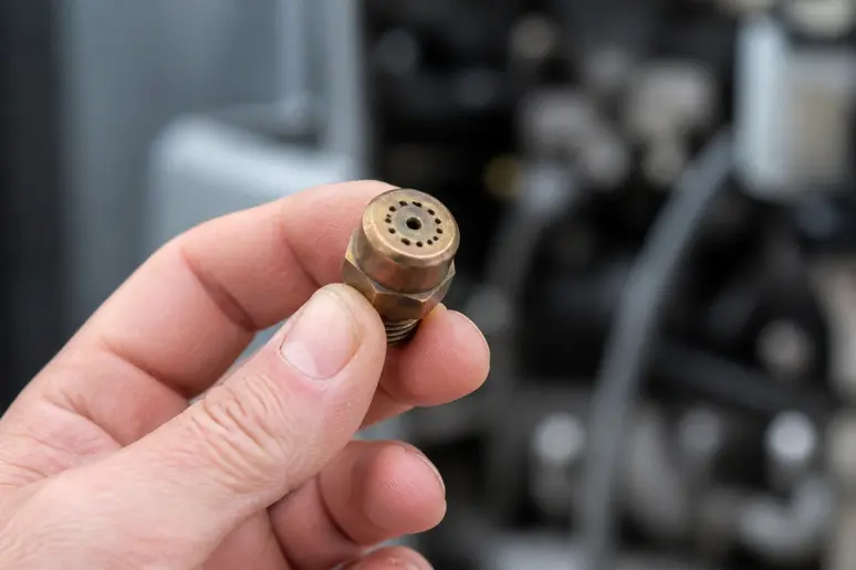Gros plan sur un gicleur de chaudière fioul en laiton, tenu par une main gantée.