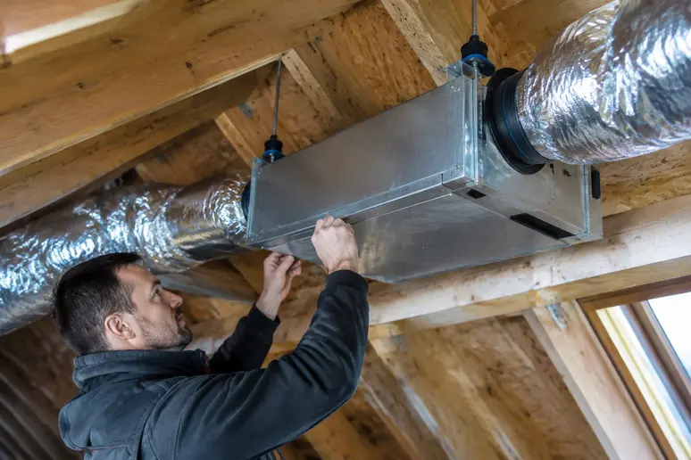 Technicien fixant un caisson de VMC sur des suspensions anti-vibratiles dans des combles.